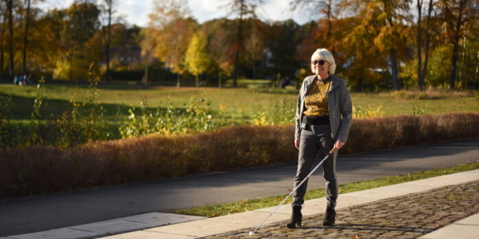 An elderly lady is walking along a street next to a meadow with a cane and a navi belt.