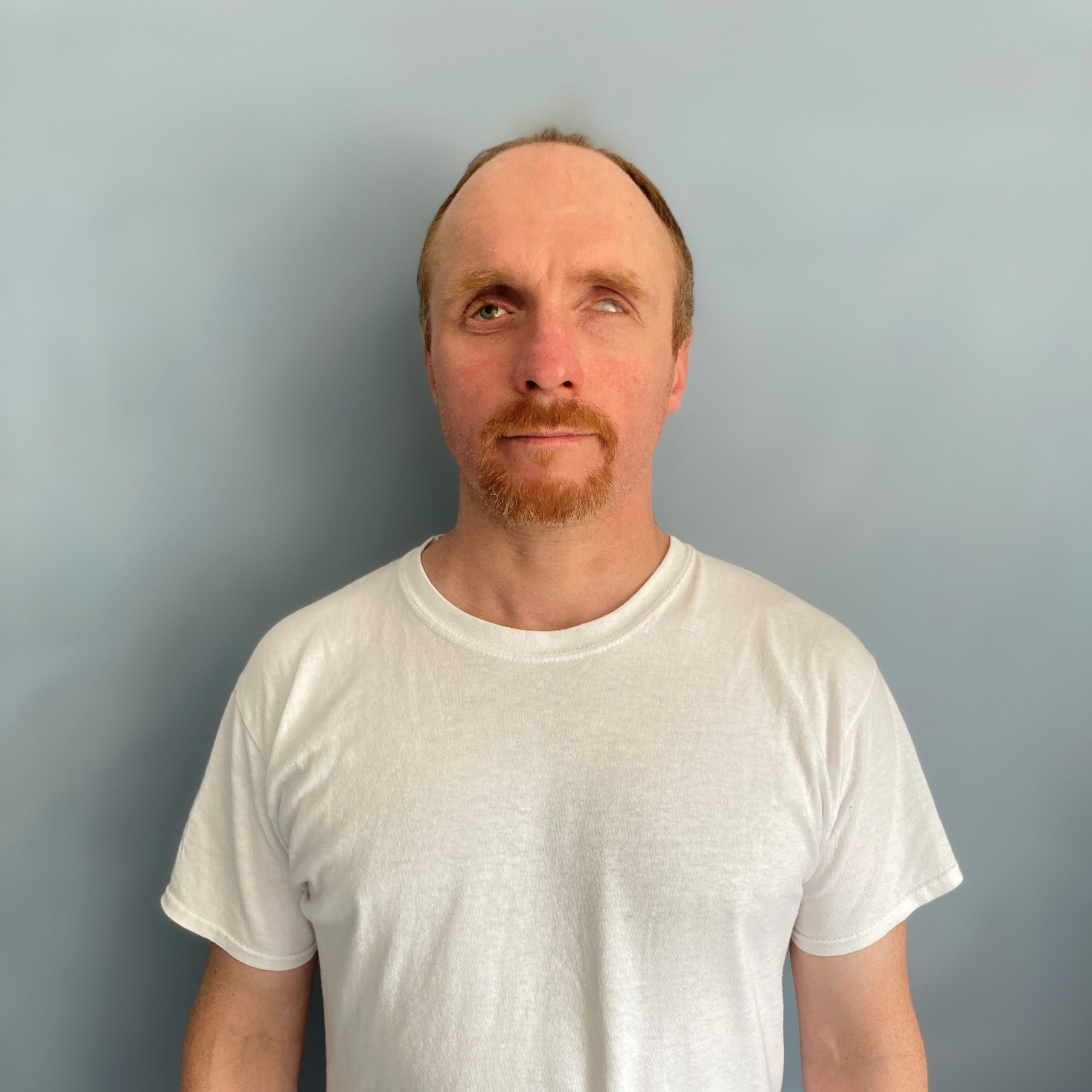 Markus Böttner, customer service, with short, reddish-brown hair, a high forehead and a reddish-brown beard, stands in a white T-shirt in front of a gray wall and looks friendly into the camera.